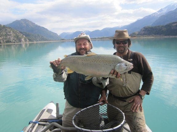 Two men proudly hold a large fish by a lake with mountains in the background.