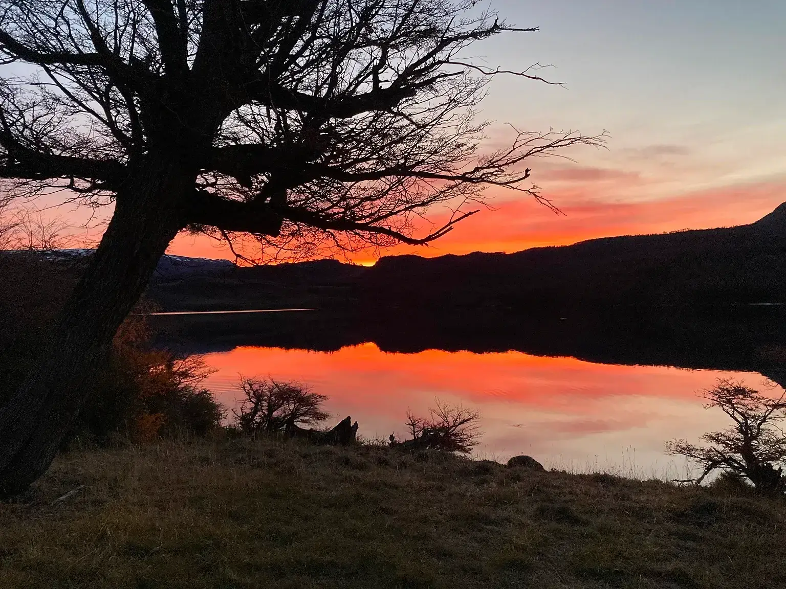A vibrant sunset over a calm lake with silhouetted trees and hills.